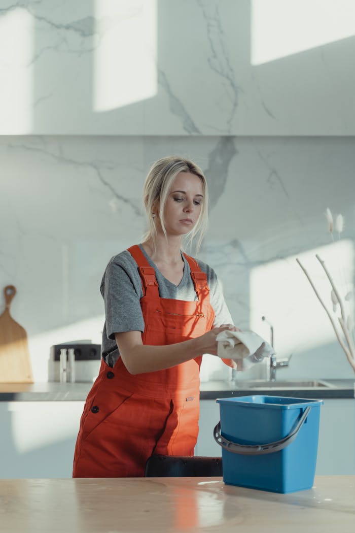 services-04 A woman wearing orange overalls cleans a kitchen counter with a rag and pail in a modern kitchen.
